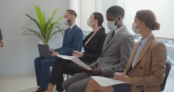 Diverse Job Candidates in Safety Mask Sitting in Office Hallway Waiting for Secretary to Invite Them