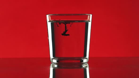 Drop of Black Ink Dripping Into Clear Water in Glass Standing on Red Background in Slowmotion