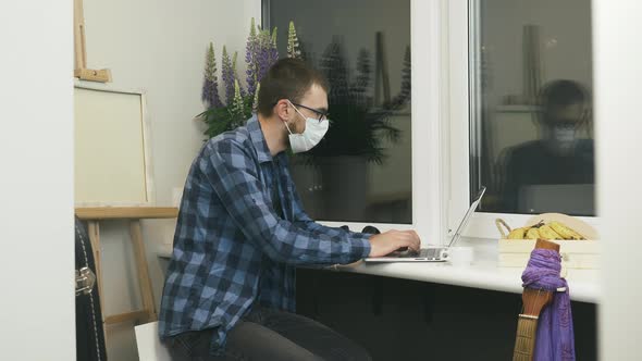 Male student in protective medical face mask using laptop computer at home workplace studying online