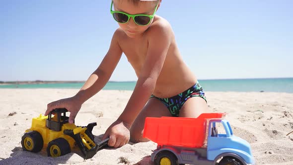 Boy Dressed in Swimming Trunks, Panama Hat and Sunglasses, Playing with Cars on a Sandy Beach