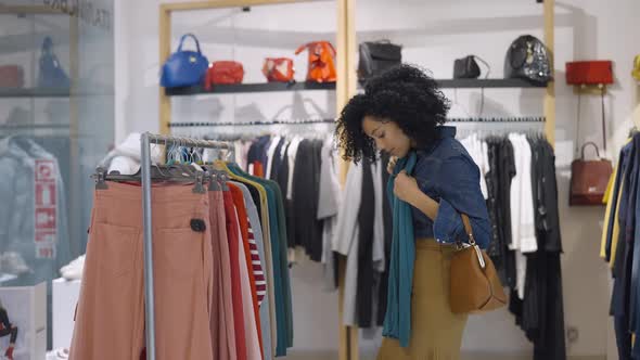 Female Customer Makes Purchases in Clothing Store Chooses Stylish Clothes Hanging on Rack Hangers