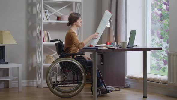 Skilled Professional Disabled Female Architect Analyzing Blueprint Sitting in Home Office Indoors