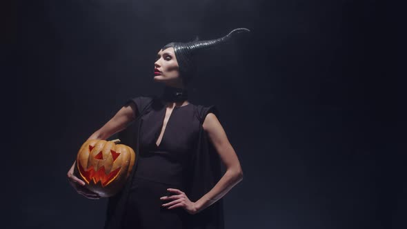 Beautiful Woman Stands on Black Background in Smoke in Sorceress Costume with Pumpkin at Hand Front