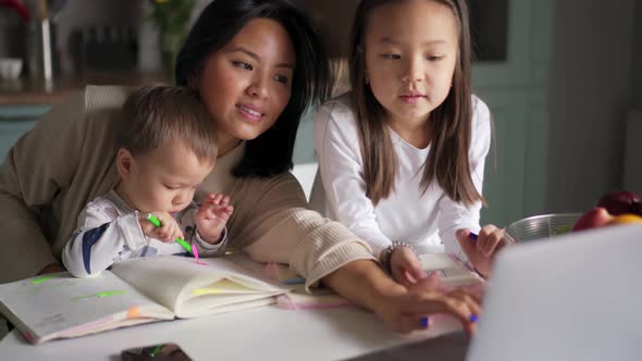 Smiling Asian mom looking at laptop with daughter while her son painting