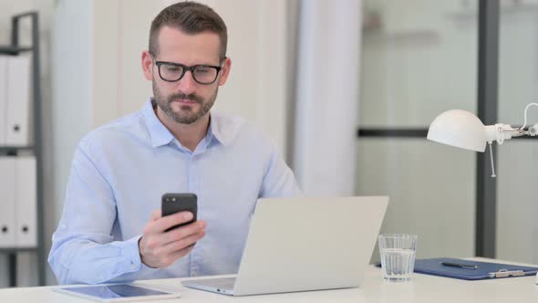 Middle Aged Man Using Smartphone While Working on Laptop