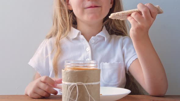 Child eating crispbread with peanut butter sitting at table home kitchen Girl with bread slice