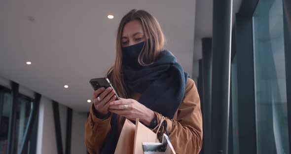 Happy Young Beautiful Caucasian Woman in Face Mask Using Smart Phone at Empty Mall Passage Carrying