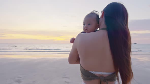 Happy mother with baby boy walks by ocean on the beach in summer