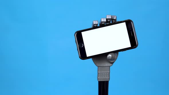 A Mechanical Hand Holds a Phone with a White Screen. Gray Cyborg Hand Holding a Smartphone on a Blue