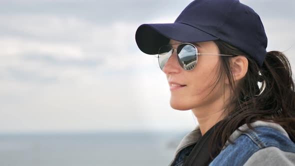 Portrait of Hiker Casual Woman in Sunglasses and Cap Enjoying Seascape at Overcast Weather