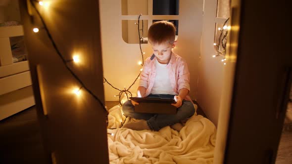 Dolly Shot of Smiling Little Boy Sitting in His Small Toy House and Using Tablet Computer