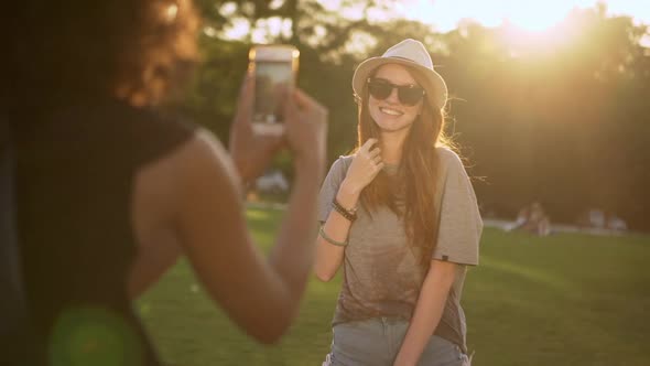 Pretty Ginger Young Girl in Hat and Sunglasses Posing While Her Tanned Friend Taking Photo of Her in