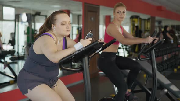 Lazy Overweight Woman Riding Sluggishly at Exercise Bike and Scrolling on Phone