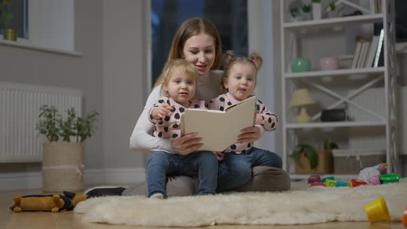 Wide Shot Portrait of Young Caucasian Mother Reading Book for Twin Daughters Sitting in Living Room