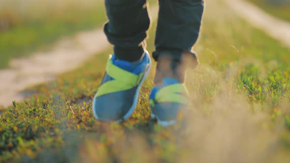 A Child in Bright Sneakers Walks on the Green Grass in the Meadow During Sunset