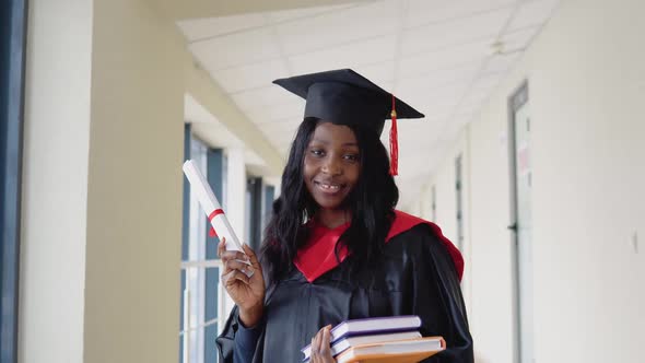 African American Female Graduate in Mantle Stands with a Diploma and Books in Her Hands and Smiles