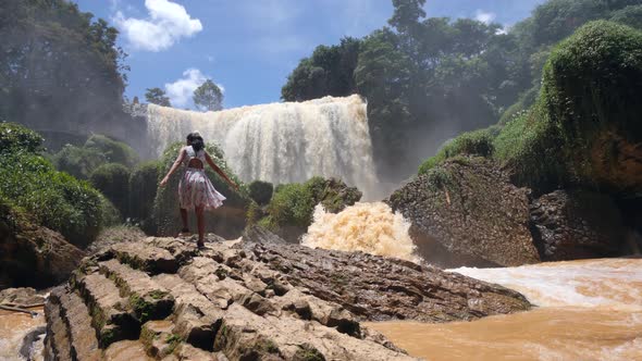 Young Woman in Floral Dress Walking on a Huge Rock Near a Massive Waterfall Flow