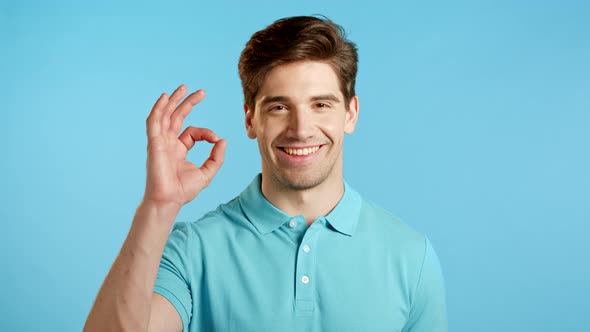 Handsome Young Man in Blue Smiles To Camera. Guy Showing OK Sign Over Minimal Background. Winner