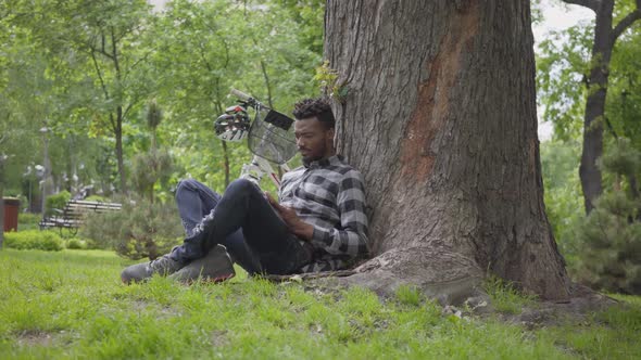 Handsome African American Man Sitting Near His Bicycle Under an Old Tree in the Park Reading a Book