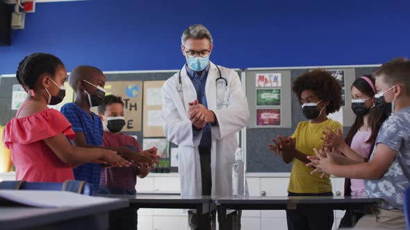 Diverse schoolteacher and schoolchildren standing disinfecting hands, all wearing face masks