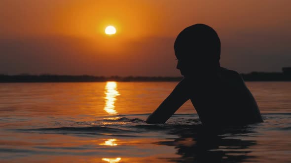 Silhouette of Boy Sitting in the Water on Background of Sunset and Orange Path. Slow Motion