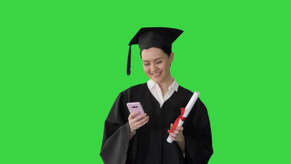 Happy Female Graduate Holding Diploma and Texting on Her Phone on a Green Screen, Chroma Key.