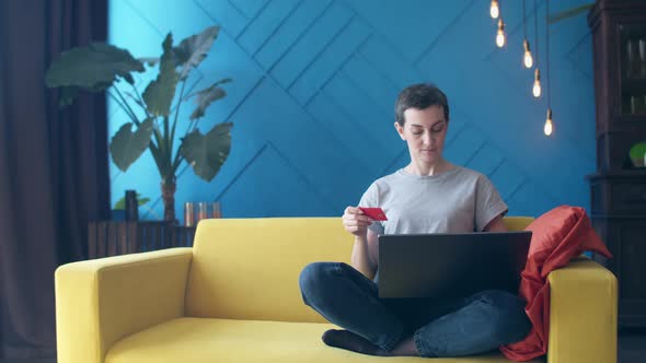 Short-haired woman enters bank card details into a laptop while sitting on the couch in a room.