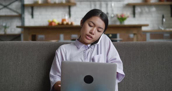 Busy Asian Business Woman Professional Talking on Phone Using Laptop Sitting on Sofa at Living Room