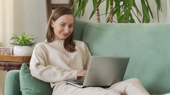 Young Brunette Woman Sitting on Couch and Doing Project on Laptop in Cozy Living Room at Home
