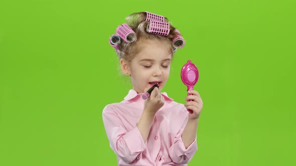 Child in Curlers Holds a Mirror in Her Hands and Paints Lipstick with Lipstick. Green Screen