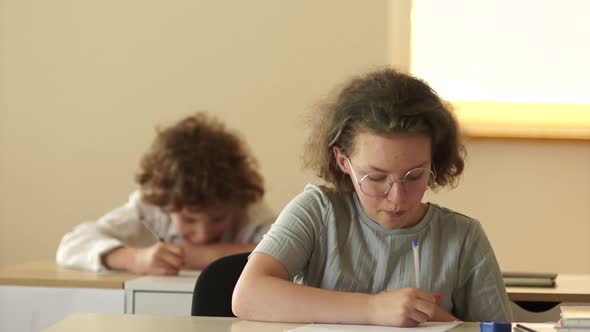 A Curlyhaired Schoolgirl with Glasses is Writing a Test and Peeping at a Cheat Sheet