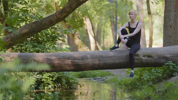 Middleaged Caucasian Woman Slim Athlete Lady in Black Tracksuit Sportwear Sits on Log Tree in Park
