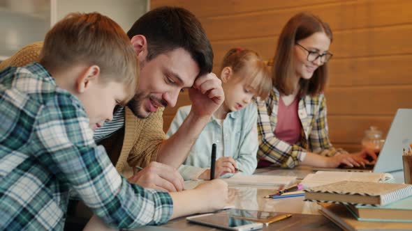 Cheeful Guy Doing Homework with Little Son Talking Laughing While Woman Teaching Daughter at Home