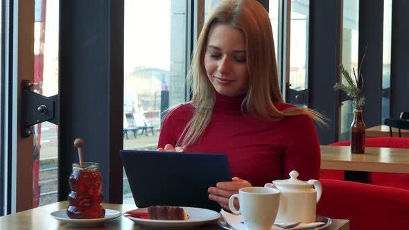 A Young Attractive Woman Sits at a Table with Meal in a Cafe and Works on a Tablet with a Smile