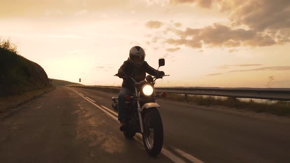 Biker Riding Motorcycle on Winding Mountain Road at Sunset with Turned on Headlight