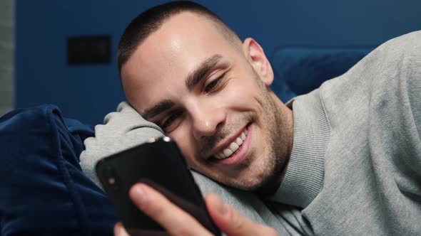 Happy Young Man Uses Smartphone While Sitting on a Sofa at Home. Man Browses Through Internet