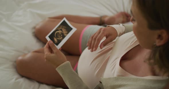 Caucasian pregnant woman sitting on bed, touching belly and looking on ultrasound photo