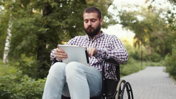 Caucasian Disabled Man is Sitting Wheelchair in Park with Tablet in His Hands and Typing Something