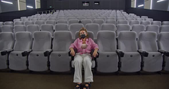 a Young Stylish Woman in a Protective Face Mask is Sitting on the First Row of an Empty Cinema Hall