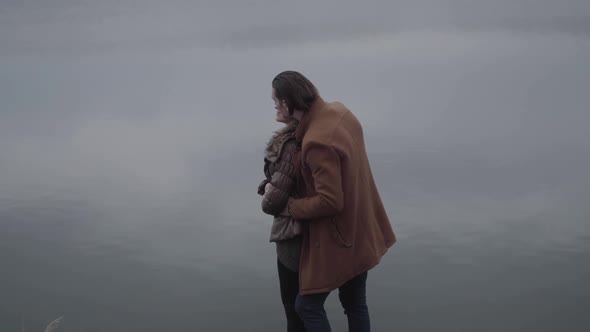 Side View of Laughing Caucasian Couple Hugging at the Background of Grey Water on Cloudy Day