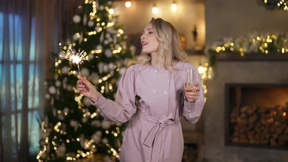 a Blonde in a Festive Dress Dances with a Glass and a Sparkler in Hands Against the Background of a