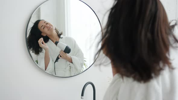 Happy Woman in Bathrobe Drying Hair with Fan and Dancing Looking at Mirror in Bathroom