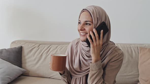 Joyous Woman in Hijab Holding Mug and Talking on Phone at Home