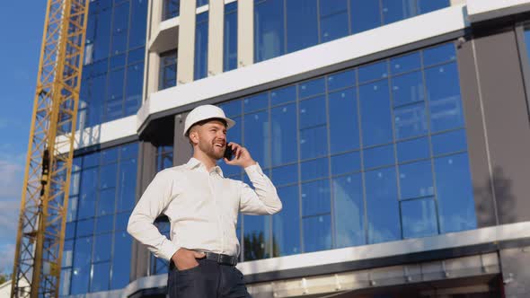 Engineer Architect in a White Shirt and Helmet on the Background of a Modern Glass Building Speaks