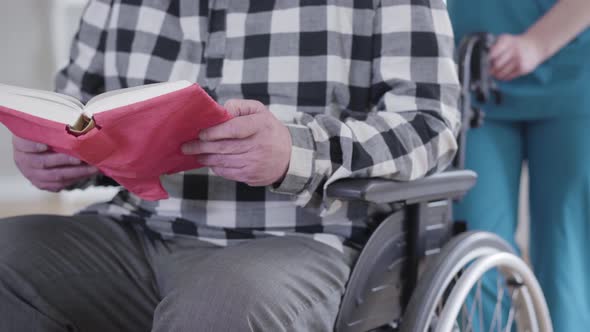 Close-up of Unrecognizable Caucasian Elderly Man Sitting in Wheelchair and Reading Book As Invalid