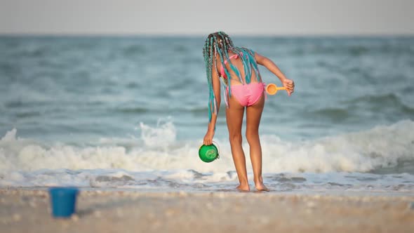 A Girl with African Braids in a Summer Costume Plays on the Beach with Shells Near the Sea with