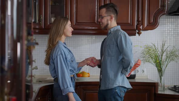 Side View of Joyful Young Man Surprising Woman with Gift on Weekend Morning in Kitchen