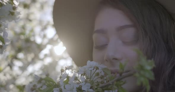 Closeup of Girl Wearing a Hat and Smelling the Flowering Branch of Apple Tree