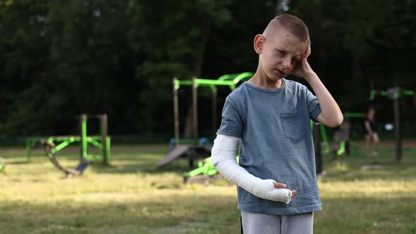 sad little boy with broken hand holding his head outdoors on summer day. 9 years child with a fractu