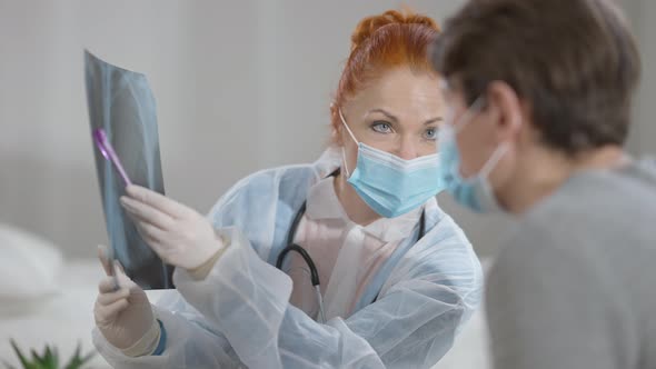 Portrait of Focused Caucasian Woman in Covid Face Mask Pointing at Lungs Xray Talking to Blurred Man